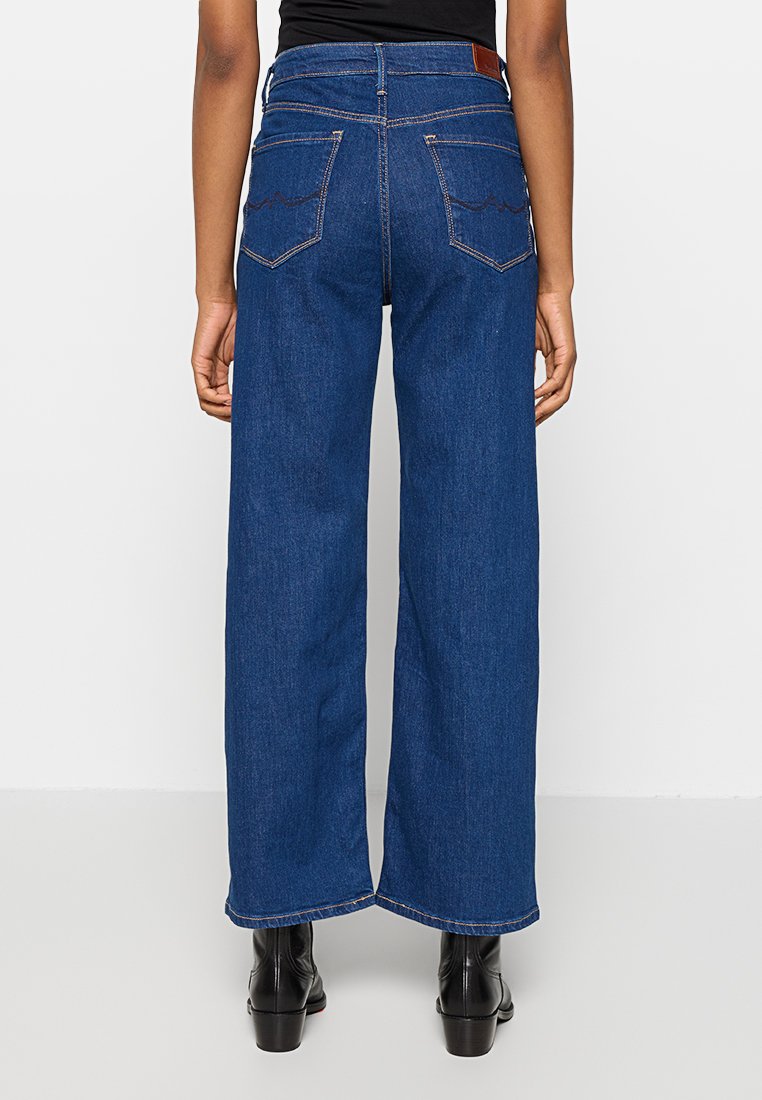 Personne portant un jean blue taille haute et large avec des bottes noires à la cheville, se tenant contre un fond blanc uni, vue de dos.
