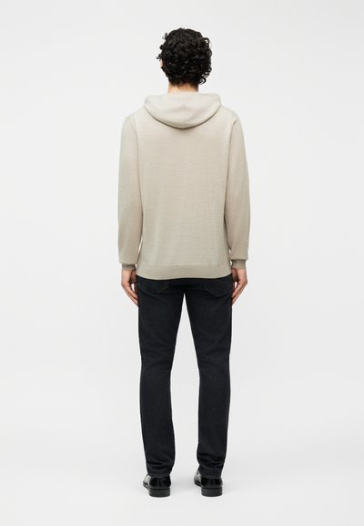 Άνδρας με σγουρά σκούρα μαλλιά, ντυμένος με ανοιχτό μπεζ φούτερ με κουκούλα, μαύρο τζιν και μαύρα παπούτσια, στεκεται με την πλάτη γυρισμένη σε μονόχρωμο φόντο.