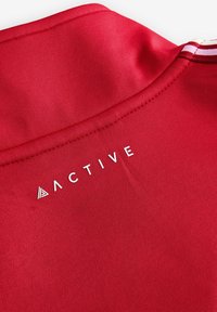 Tissu de sport rouge avec gros plan sur la couture du col et le texte blanc "ACTIVE" accompagné d'un symbole triangulaire près de l'encolure.