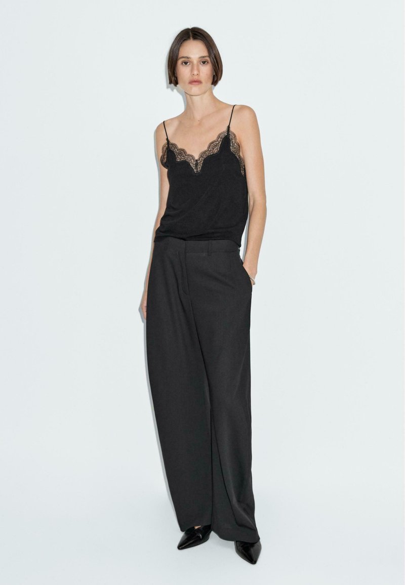 Camisole noire avec des détails en dentelle associée à un pantalon large taille haute noir. Tissu lisse, lignes épurées et talons aiguilles noirs.