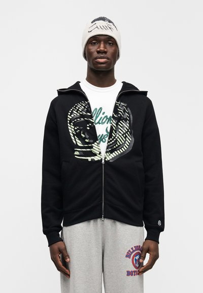 Μαύρο φούτερ με φερμουάρ και μεγάλο γραφικό σχέδιο σε ανοιχτό πράσινο, συνδυασμένο με λευκό μπλουζάκι και γκρι φόρμες, ολοκληρωμένο με πλεκτό καπέλο.