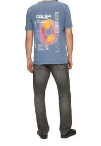 Blauwe katoenen t-shirt met grafische print met een cirkelontwerp en tekst op de achterkant. Gepaard met donkergrijze spijkerbroek en zwarte schoenen.