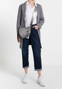 Harmaa crossbody-laukku, jossa on sileä ja matta pinta, pyöreä muoto ja säädettävä olkahihna, käytetty tarkastetun takin ja farkkujen kanssa.