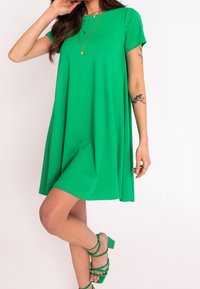 Grünes kurzes Kleid mit lockerer Passform, aus weichem Stoff. Kombiniert mit grünen Riemchen-Absätzen, hat eine glatte Textur und einen runden Ausschnitt.