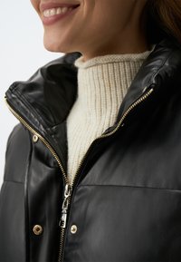 Veste en cuir noir avec un col haut, finition brillante et quincaillerie dorée. Présente un pull crème à côtes en dessous.