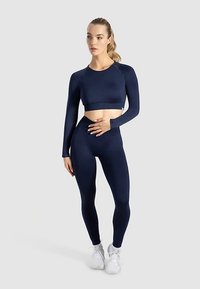Marineblaue, eng anliegende Sport-Crop-Top und Leggings aus dehnbarem Material, mit langen Ärmeln und nahtlosem Design. Weiße Sneakers runden das Outfit ab.