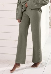 Olivgrüne, weit geschnittene Hose aus weichem Material. Verfügt über ein Logo am Unterschenkel und gerippte Bündchen. Auf einer strukturierten weißen Fläche platziert.