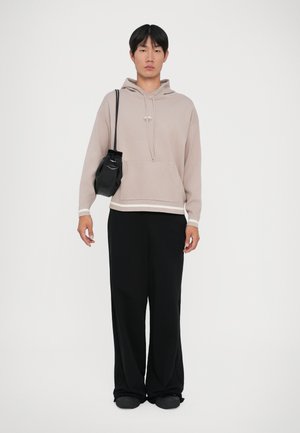 Giovane con felpa beige, pantaloni neri a gamba larga, scarpe nere e che porta una piccola borsa a tracolla nera su uno sfondo bianco.