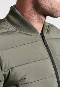 Chaqueta acolchada verde oliva con cuello acanalado y cremallera negra. Presenta detalles de costura horizontal, textura suave y material liviano.