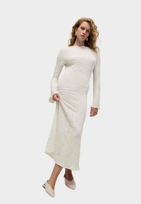 Robe blanche ajustée à manches longues en tissu texturé, coupe au niveau du genou, avec des détails subtils de couture, assortie à des chaussures à talons beiges.