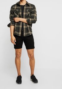 Hombre que lleva zapatillas negras, pantalones cortos negros, camiseta negra y una camisa de cuadros verde y negra sin abotonar, de pie contra un fondo blanco liso.