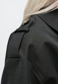 Črni plašč s tuško teksturo, z ramenskim epauletom in dekorativnim gumbom. Gladek material z minimalnimi detajli.