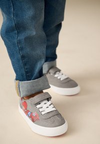 Baskets en toile grise avec semelles en caoutchouc blanches, présentant un graphisme rouge de Spider-Man sur le côté. Portées avec un jean bleu retroussé.