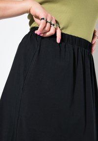Zwarte rok met elastische tailleband, gemaakt van zachte stof. Hand die een zak vasthoudt, met details zoals plooien en een gladde textuur.