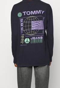 Svart långärmad skjorta med grafiska mönster i lila och grönt, inklusive en glob, textelement och mönster. Avslappnad passform med ribbade ärmslut.