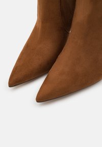 Botas marrones de ante con punta afilada y textura suave, mostradas en primer plano sobre un fondo blanco.