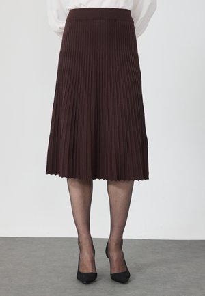 Femme portant une jupe midi plissée marron foncé, des bas noirs transparents et des escarpins noirs à bout pointu, debout sur un sol gris.