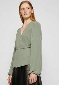 Green textured wrap top with long puff sleeves and a deep V-neck. Paired with a black leather skirt, highlighting the top's flowing design.