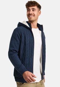 Navy blue zip-up hoodie with a textured cable knit design, fleece-lined interior, and ribbed cuffs. Model wearing a white shirt underneath.