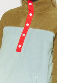 Polarowy sweter z wysokim kołnierzem; miętowozielona baza i brązowe rękawy; wyposażony w czerwoną listwę z białymi guzikami; ma kieszeń z przodu.