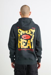 Grå hættejakke med en iøjnefaldende, gul grafik, der står "SWEET HEAT" med røde accenter og et Nike-logo; blød tekstur og afslappet pasform.
