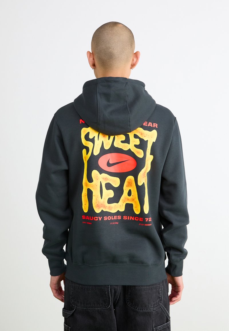 Grå hættejakke med en iøjnefaldende, gul grafik, der står "SWEET HEAT" med røde accenter og et Nike-logo; blød tekstur og afslappet pasform.