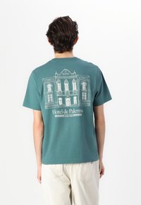Camiseta de algodón color verde azulado con un diseño ilustrado en blanco de un edificio y el texto "Hotel de Palermo" en la parte posterior. Cuello redondo, mangas cortas.