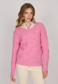 Pull en tricot rose avec un motif texturé, à col rond et à manches longues, porté sur une chemise blanche à rayures claires.