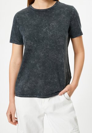 Kobieta ubrana w wyblakły czarny t-shirt z krótkim rękawem i białe spodnie z ręką w kieszeni na jednolitym białym tle.