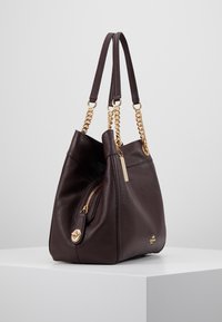 Ciemnobrązowa skórzana torebka o miękkim kształcie, zdobiona złotym łańcuszkiem i zapinana na zamek błyskawiczny. Charakteryzuje się gładką fakturą oraz detalem z logo.