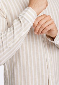 Camicia button-up di un beige chiaro con strisce verticali bianche, dotata di un polsino con bottone, realizzata in un tessuto testurizzato.
