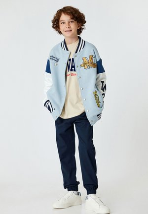 Lichtblauwe varsityjack met marineblauwe mouwen, geborduurde details en ribgebreide manchetten. Een beige t-shirt en marineblauwe joggingsbroek maken de outfit compleet. Witte sneakers.