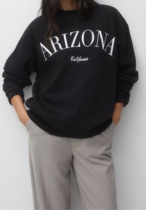 Personne portant un sweat-shirt noir avec le texte « ARIZONA California » et un pantalon gris clair, debout, les mains dans les poches, contre un mur blanc.