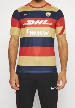 Mężczyzna w koszulce piłkarskiej z poziomymi paskami w kolorach czerwonym, granatowym i złotym z logo DHL i Mifel oraz w czarnych spodniach Nike z białym logo Swoosh.