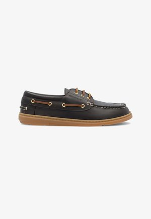 Scarpa da barca in pelle nera con suola in gomma color cuoio, lacci in pelle marrone passati attraverso occhielli dorati e dettaglio di cuciture bianche lungo la punta.