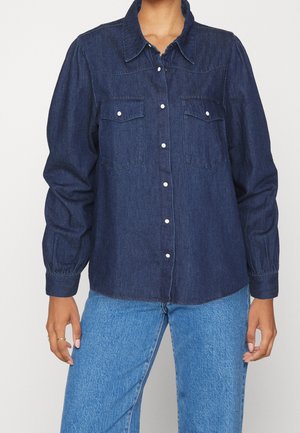 Long-sleeve dark denim button-up shirt with collar and two front chest pockets, worn with light blue jeans.