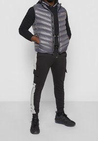 Nafouknutá šedá vesta přes černou mikinu s kapucí, v kombinaci s černými jogger pants se šedými pruhy po stranách. Oblečené s černými sportovními botami.