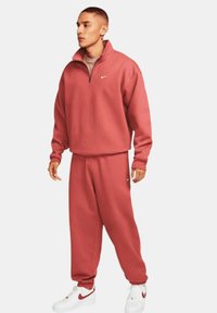 Sudadera roja de cuello alto con media cremallera y el logo de Nike, combinada con pantalones de chándal rojos a juego. Zapatillas blancas con detalles rojos.