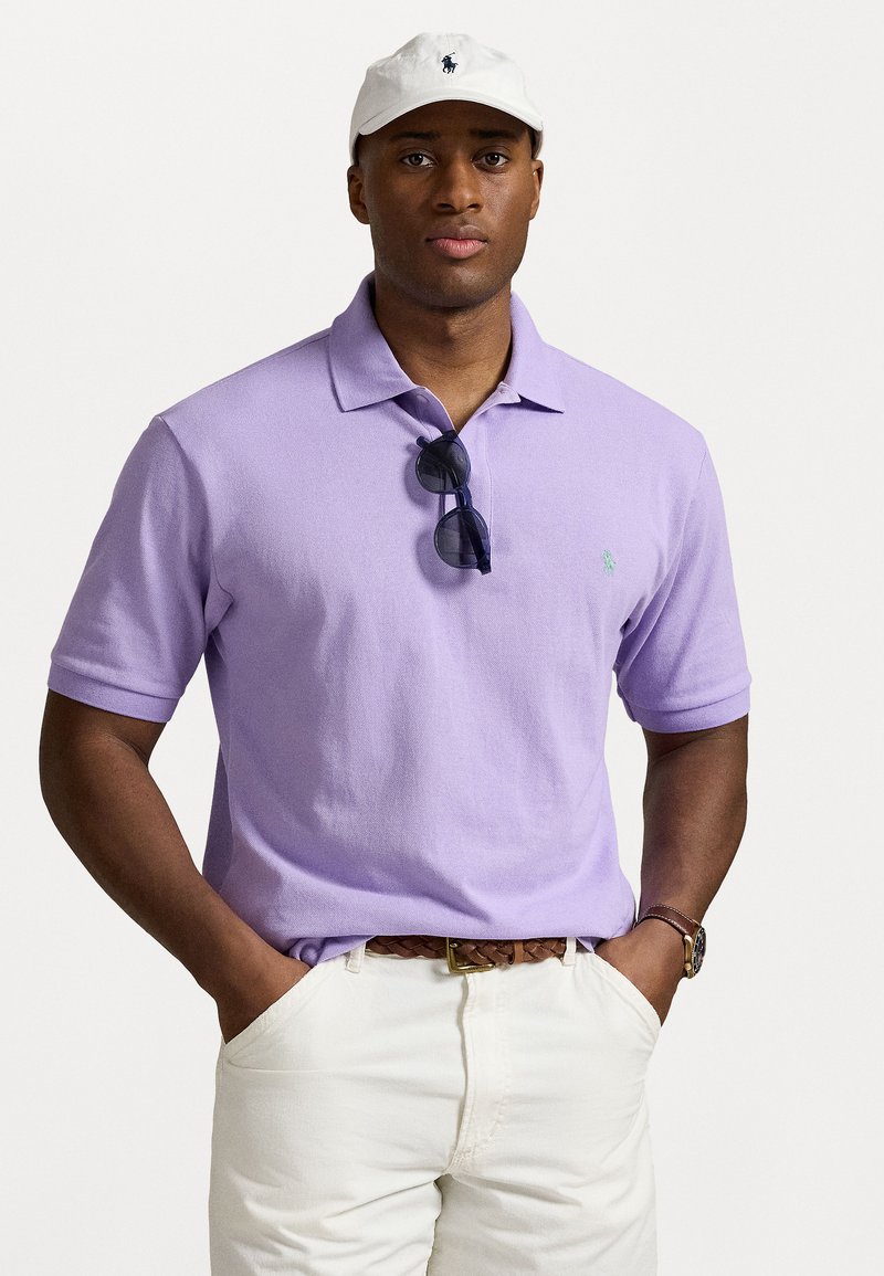Gaiši violets polo krekls ar apkakli, nēsāts virs baltām biksēm. Krekls ir ar īsām piedurknēm un mazu zaļu logotipu. Tumši saulesbrilles balstās uz apkakles.