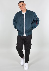 Zelená bomber bunda se zipem vepředu a žebrovanými manžetami, zkombinovaná s černými cargo kalhotami a bílými teniskami. Hladká tkanina a praktický design.