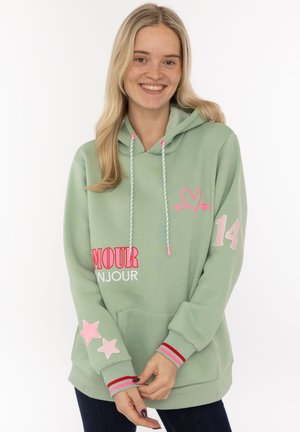 Lächelnde Frau in einem hellgrünen Hoodie mit rosa Herz, der Nummer 14, Sternen und dem Schriftzug "amour bonjour", stehend vor weißem Hintergrund.