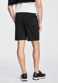 Czarne sportowe szorty wykonane z miękkiego materiału, z tylną kieszenią i luźnym krojem, w połączeniu z czarnymi butami i białymi podeszwami.