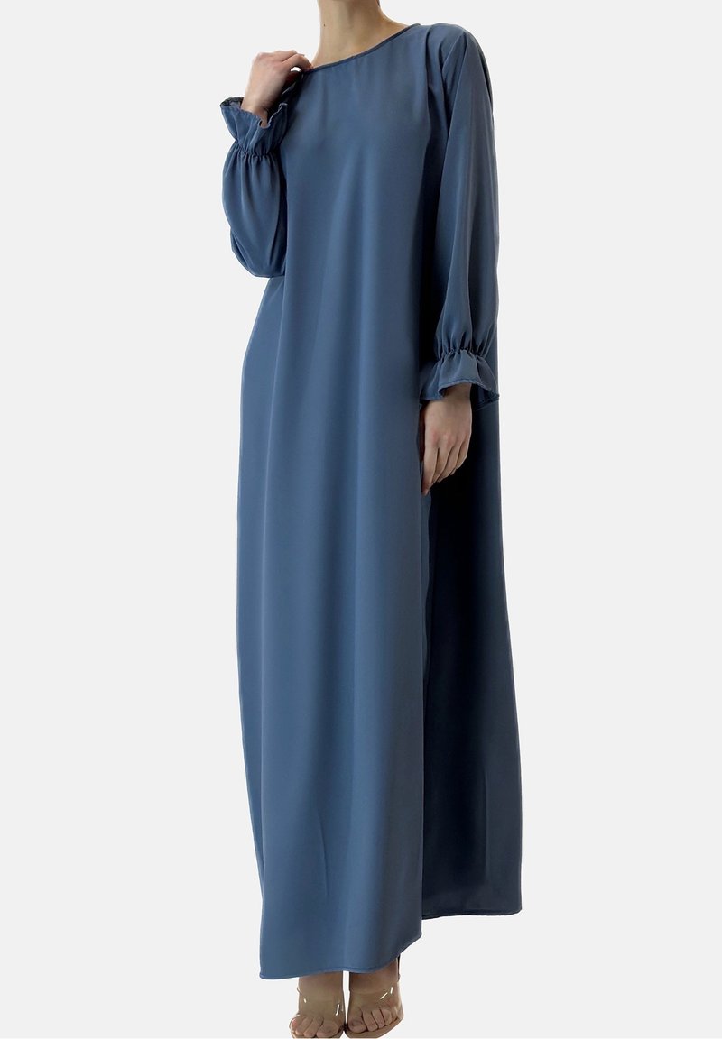 Personne portant une longue robe bleue ample avec des manches longues resserrées aux poignets, debout pieds nus devant un fond uni.