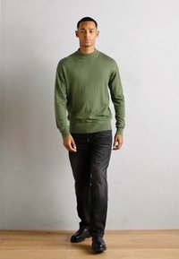 Pulover tricotat de culoare verde măsliniu cu guler înalt, purtat cu blugi negri și pantofi negri, modelat de o persoană pe un fundal neutru.