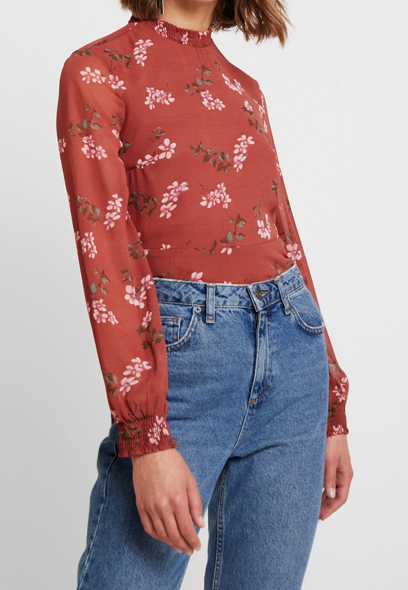 Kvinde iført en rustfarvet langærmet bluse med blomsterprint, smockede manchetter og højtaljede blå jeans, stående mod en ensfarvet baggrund.