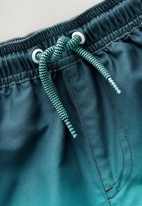 Mørkegrønne badeshorts med elastisk talje, parret med en tyk turkis snor og synlige sømdetaljer.