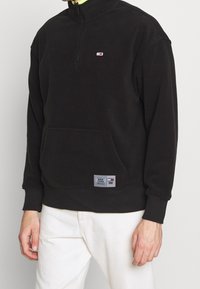Svart zip-up sweatshirt med hög krage, med en framficka och en liten logotyp på bröstet. Texturerat tyg med ribbade muddarna.