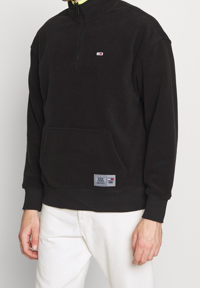 Svart zip-up sweatshirt med hög krage, med en framficka och en liten logotyp på bröstet. Texturerat tyg med ribbade muddarna.
