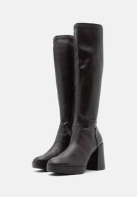 Bottes en cuir marron montant jusqu'au genou avec un talon épais, dotées d'une fermeture éclair latérale et d'une texture lisse. Bout plat et détails de couture minimalistes.