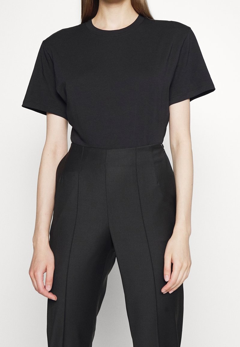 Personne portant un T-shirt noir uni à manches courtes rentré dans un pantalon noir tailleur taille haute sur fond blanc.